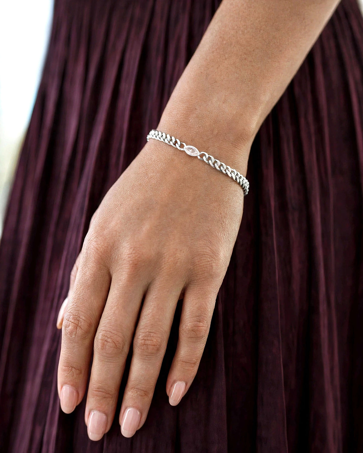 A close-up of a person's hand and wrist wearing a silver chain bracelet, set against a dark plum pleated dress.
