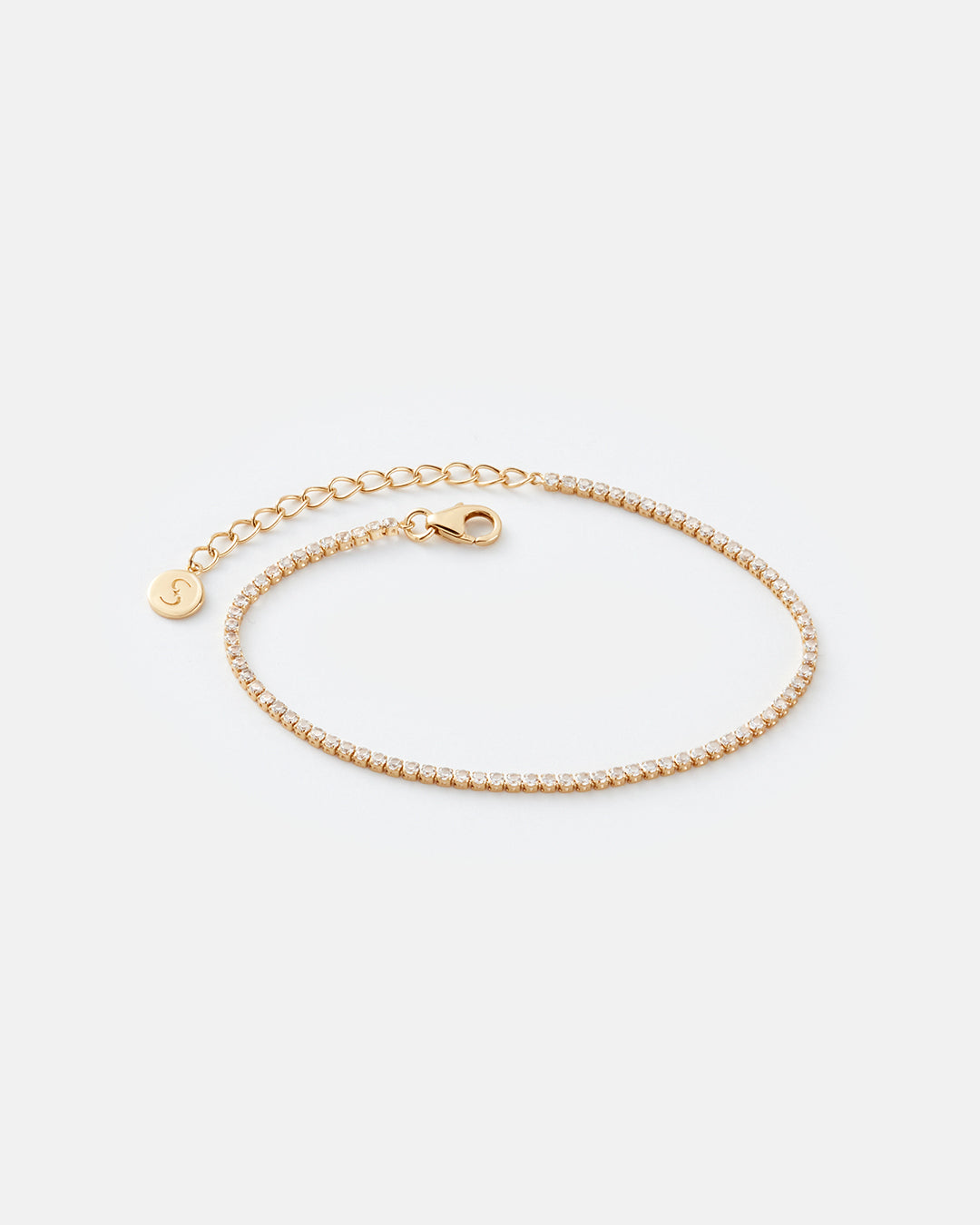A delicate gold chain bracelet with a tennis chain made of white topaz, a lobster clasp, an extender chain, and a small circular charm engraved with a stylized "S" letter, displayed on a light gray background.