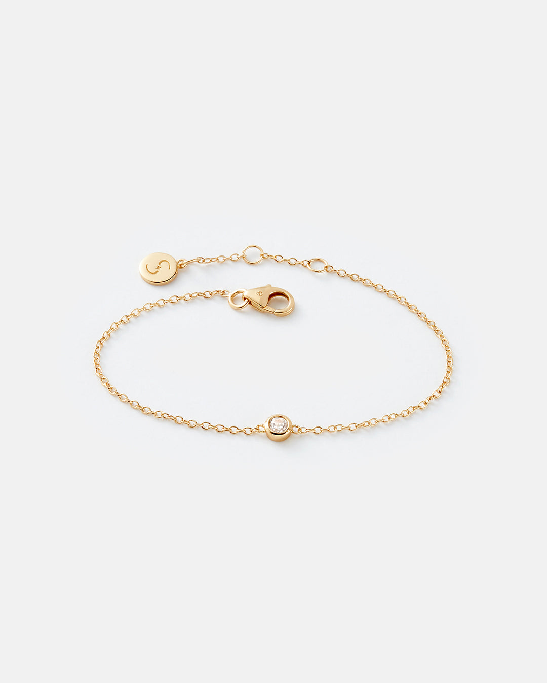 A delicate gold chain bracelet with a lobster clasp, an extender, a small round disc charm engraved with a symbol, and a central bezel-set clear white topaz gemstone, shown on a white background.