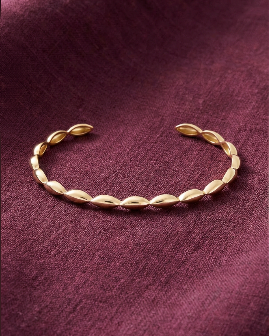 A delicate gold leaf-patterned cuff bracelet resting on a textured, deep burgundy fabric background.