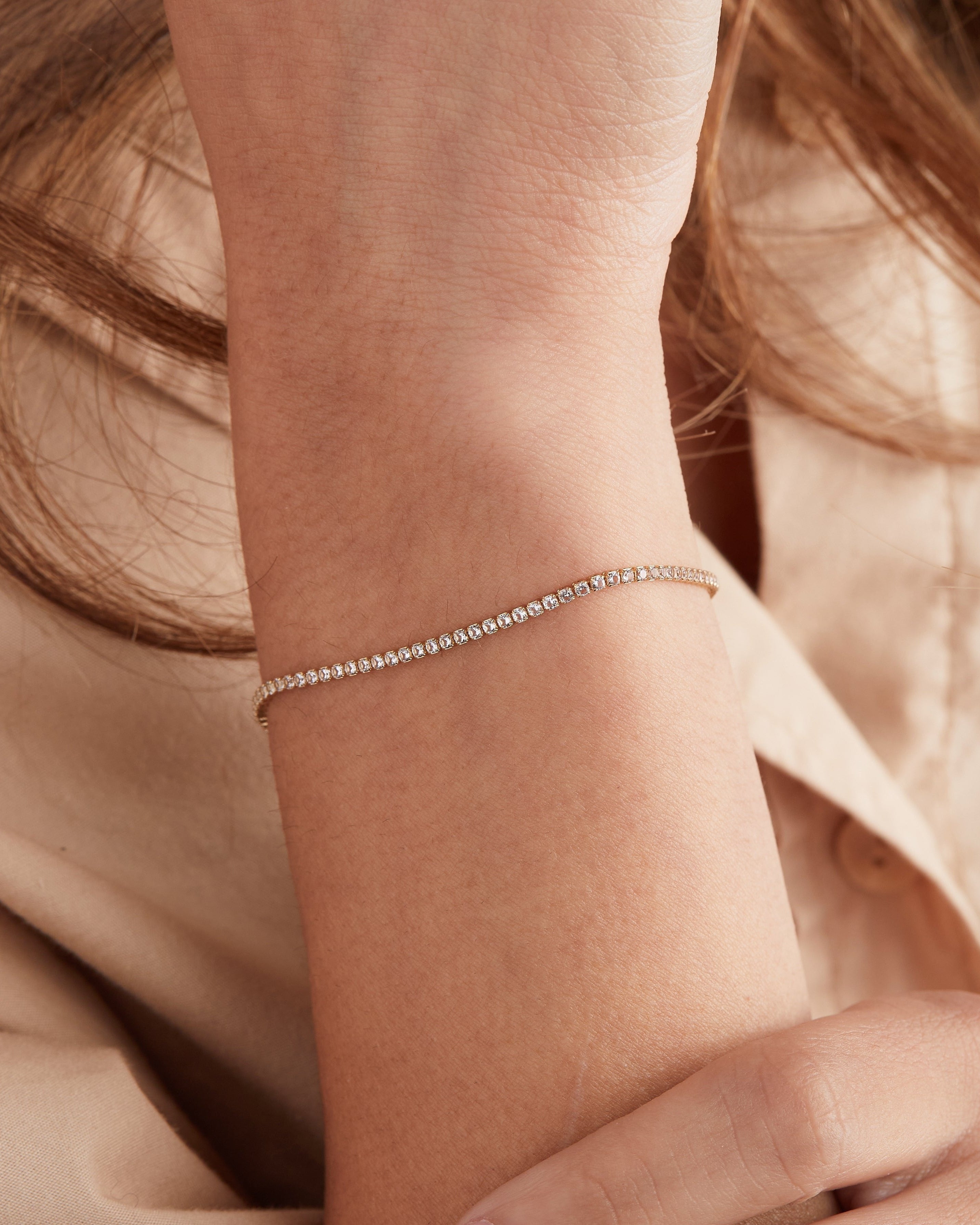 A delicate silver tennis bracelet with small clear stones worn on a person's wrist, shown against a soft beige shirt and warm neutral background.