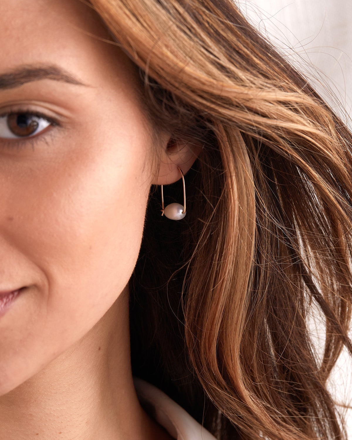 A close-up of a woman wearing a delicate gold hoop earring with a small white pearl drop, shown against her ear and brown hair.