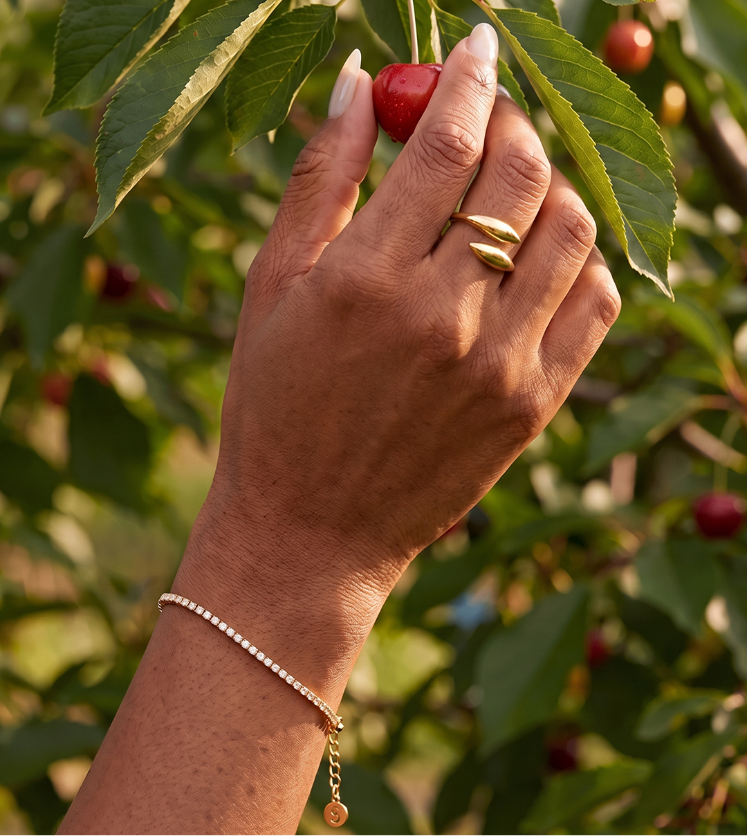 A close-up lifestyle shot of a person's hand wearing a delicate gold beaded bracelet and a gold ring, gently touching a red cherry on a tree branch with green leaves in the background.