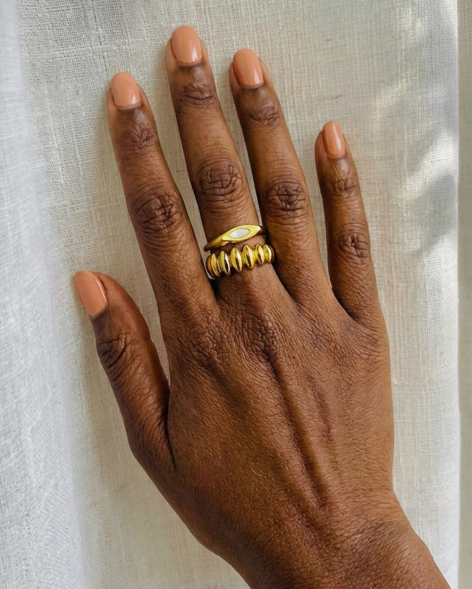 A close-up of a person's hand resting against a white curtain, wearing a textured gold ring with a small oval yellow stone on the ring finger.