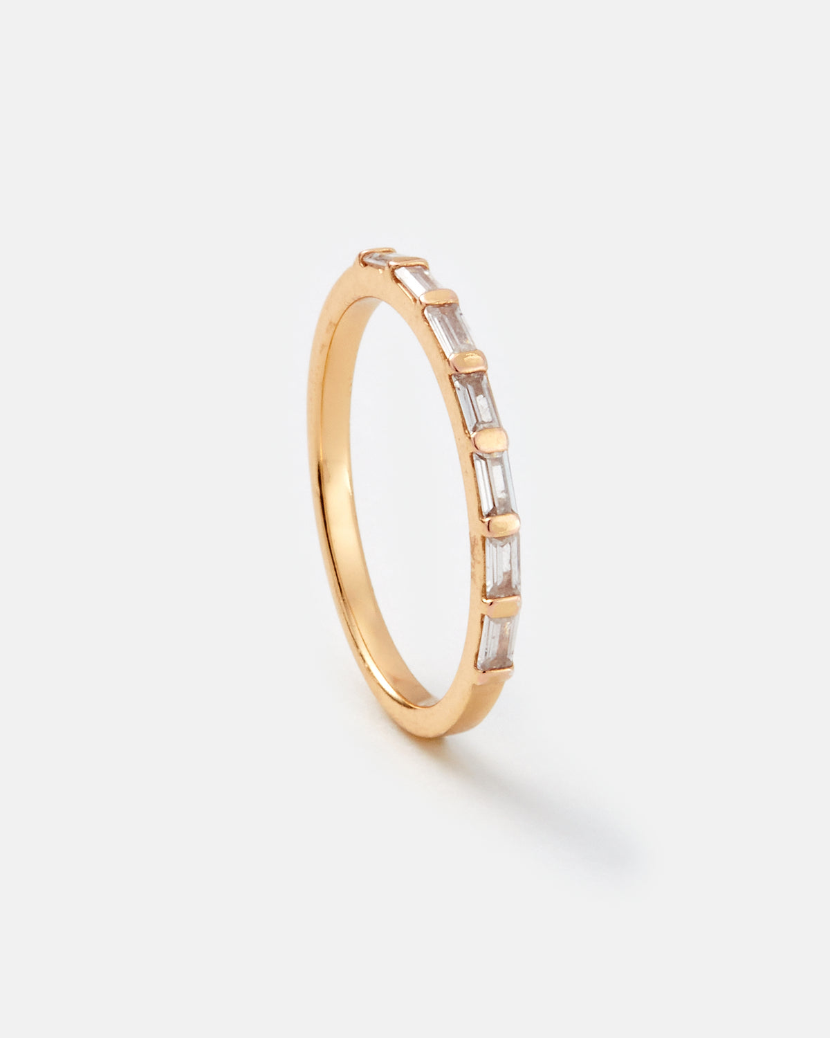 A slim yellow gold ring featuring a row of baguette-cut clear stones, photographed on a clean, light gray background.