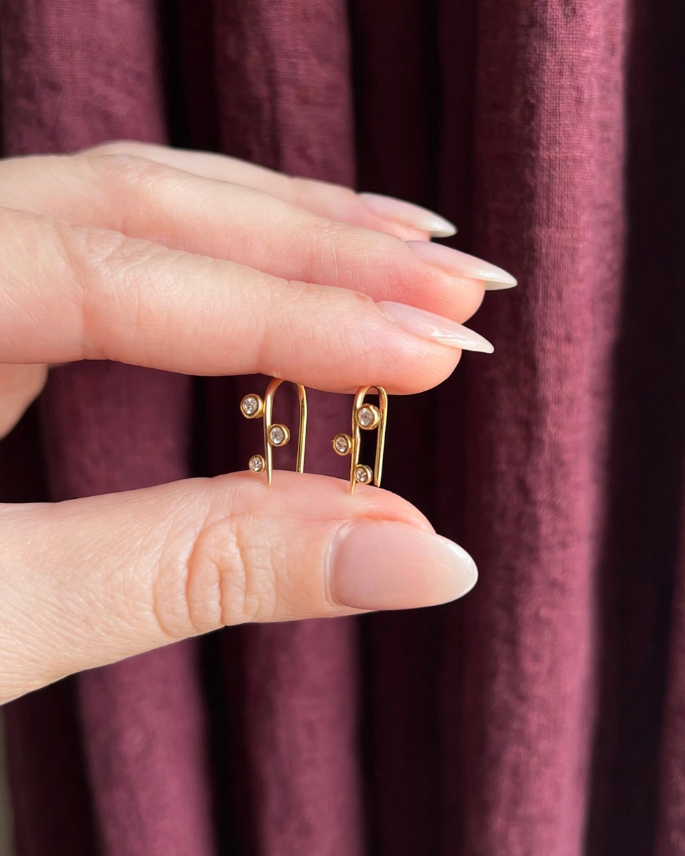 A close-up of a hand holding a pair of small gold hoop earrings with tiny clear stones, set against a dark burgundy textured curtain background.