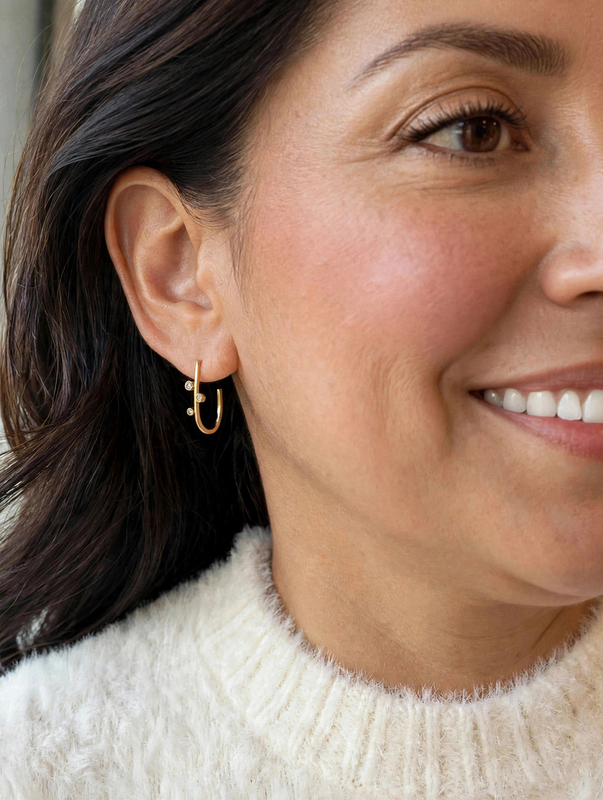 A close-up of a woman wearing a small gold huggie hoop earring with tiny dangling charms, paired with a cream-colored knit sweater.