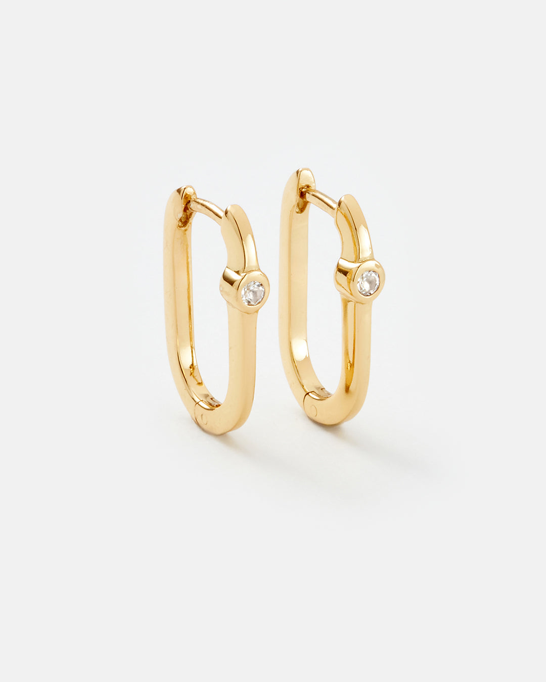 A pair of gold huggie hoop earrings with a small clear round gemstone set on the front of each earring, shown on a light gray background.