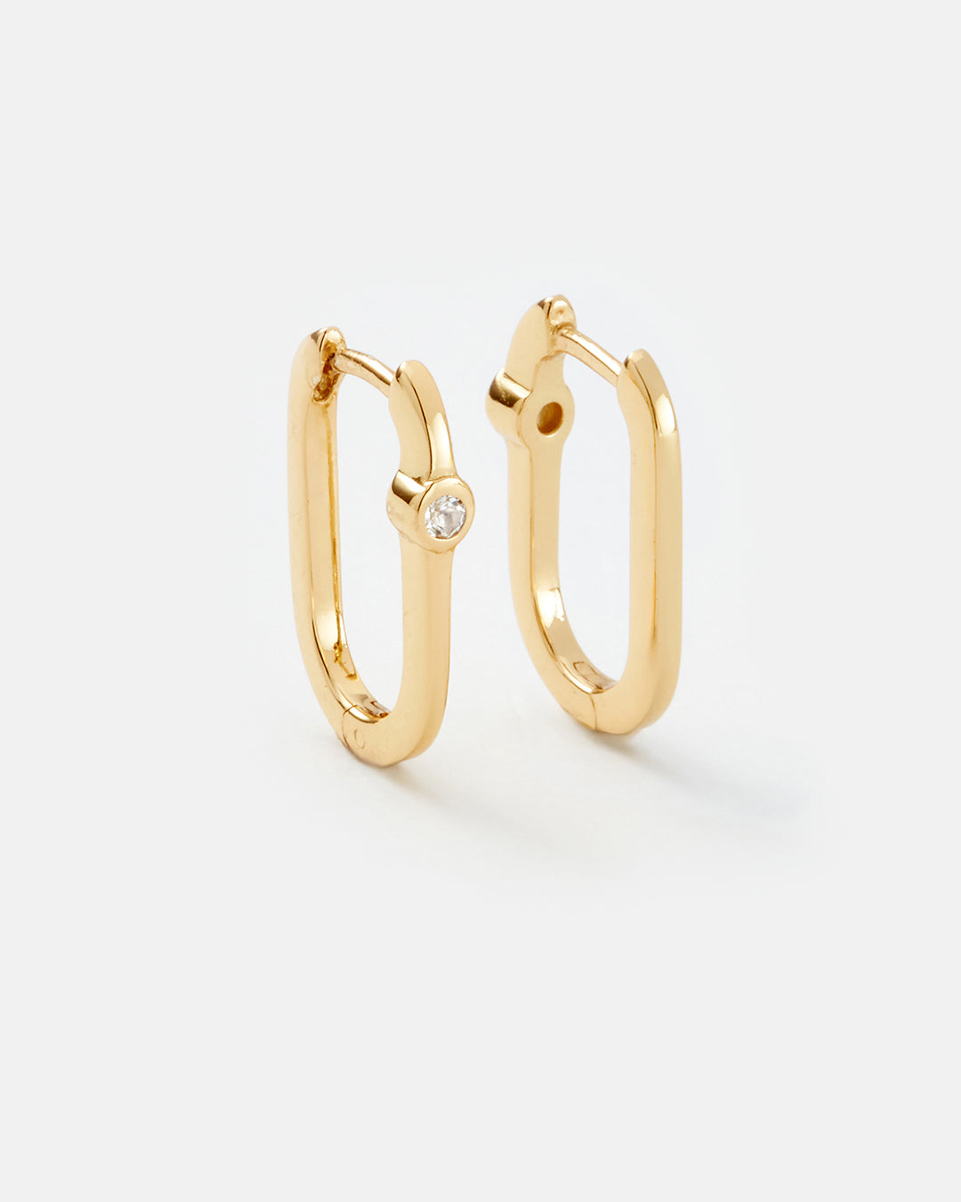 A pair of gold huggie hoop earrings with a small clear gemstone accent on one earring, displayed on a clean light gray background.
