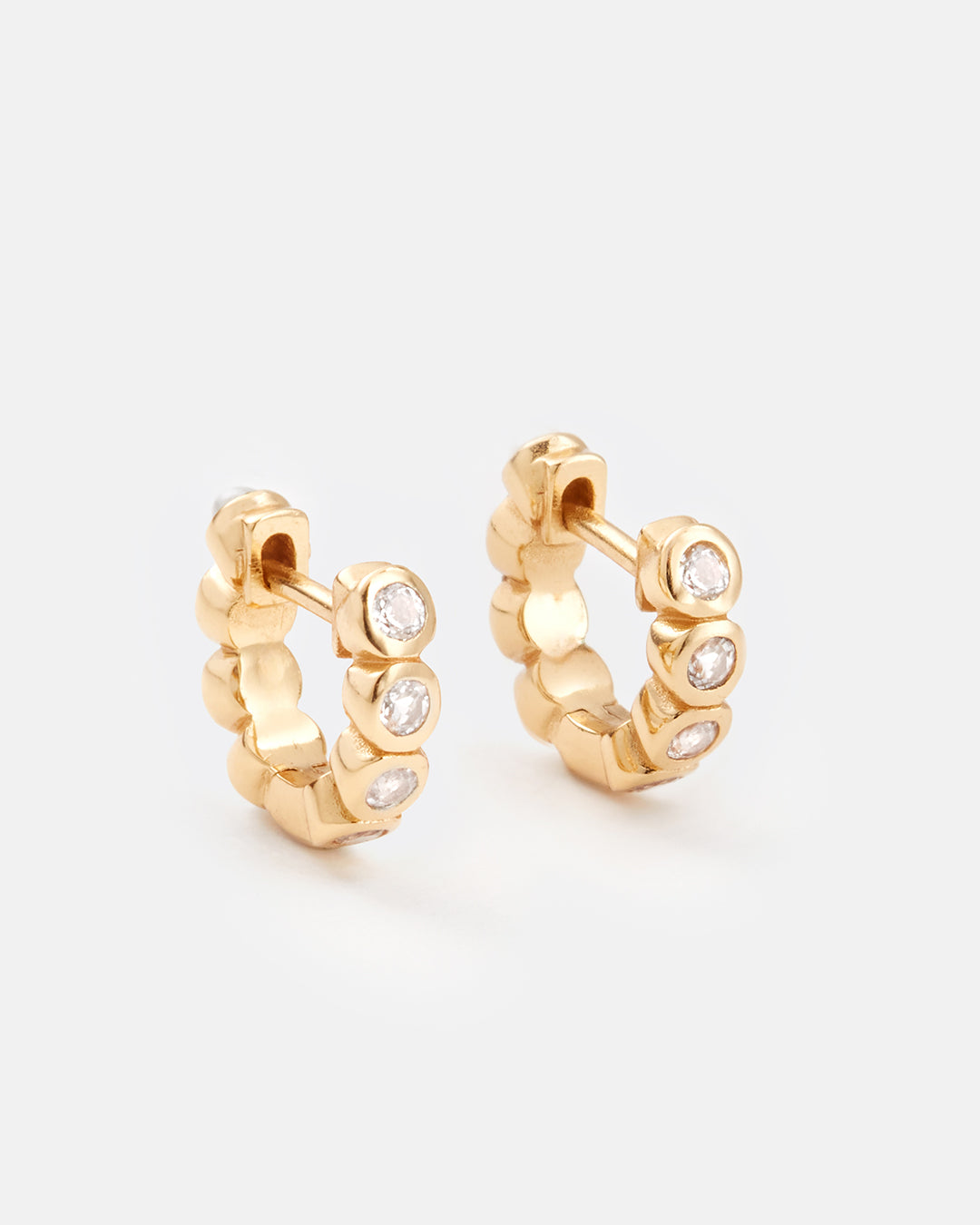 A pair of gold huggie hoop earrings with small clear round-cut stones set along the front, photographed on a clean light gray background.