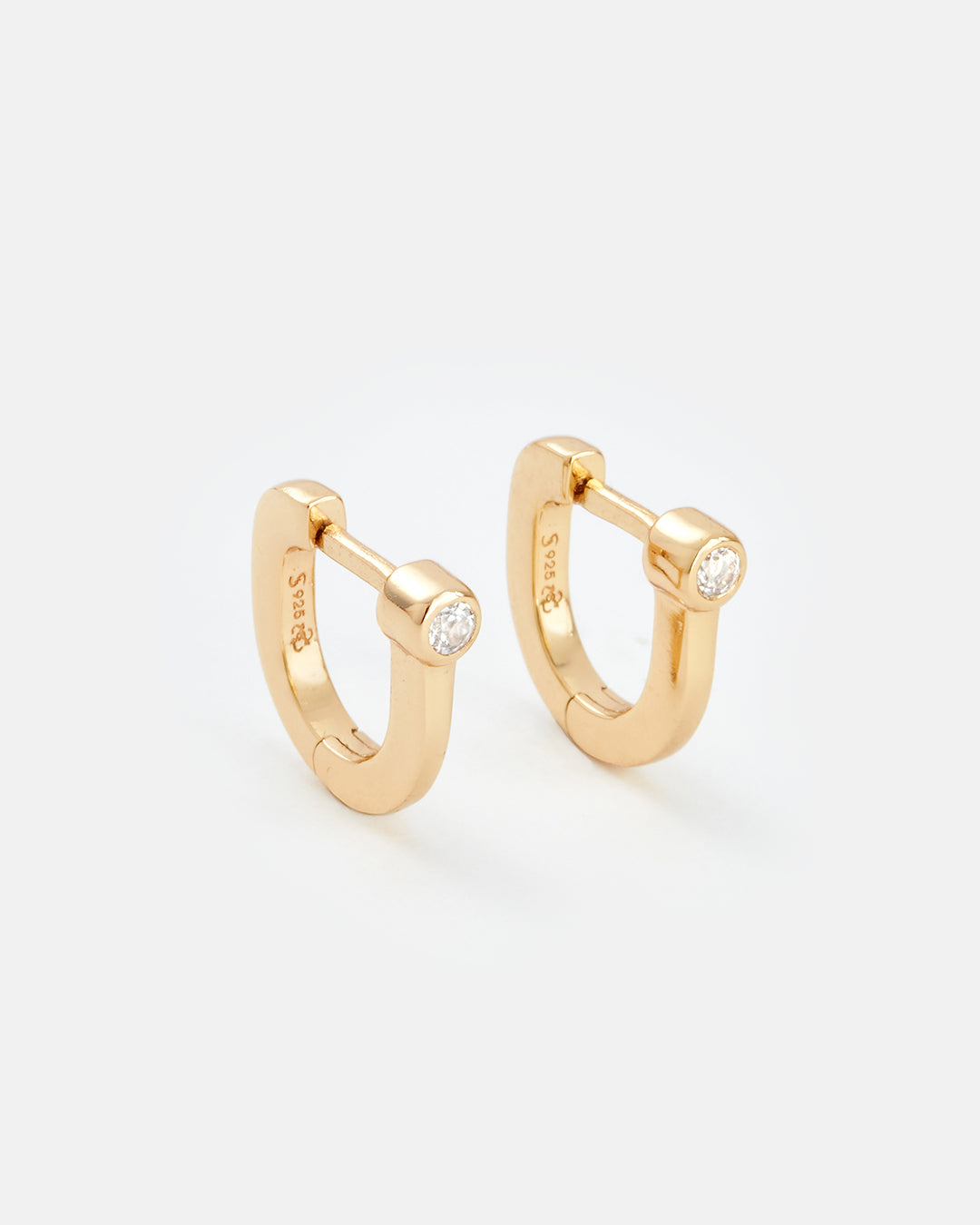 A pair of gold huggie hoop earrings, each featuring a small clear round-cut gemstone set in a bezel-like frame. The earrings are photographed on a clean, light gray background with soft shadows.