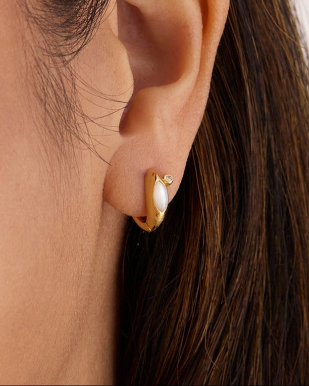 A close-up of a person's ear wearing a gold huggie hoop earring with a white faux-pearl inlay and a small clear stone.
