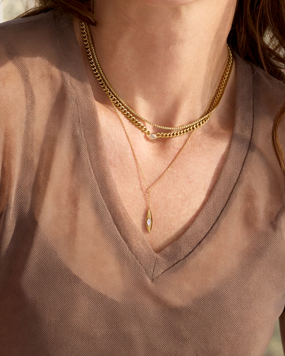 A woman wearing layered gold necklaces and small gold hoop earrings, photographed in a softly lit indoor setting with a sheer taupe-brown V-neck top.