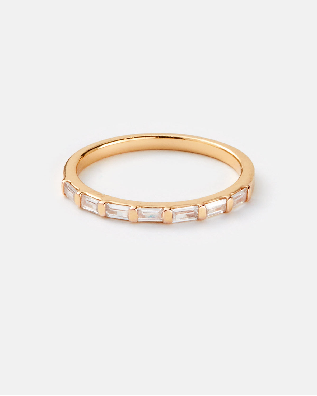 A gold band ring featuring a row of clear baguette-cut gemstones, photographed on a clean, light gray background.