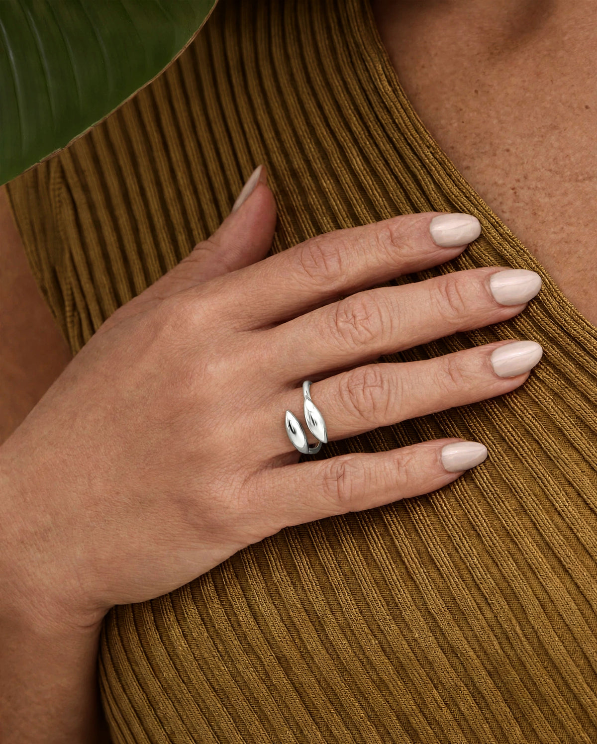 A close-up of a person's hand resting on a ribbed olive-brown top, wearing a silver wrap-style ring with two leaf-like curved bands.
