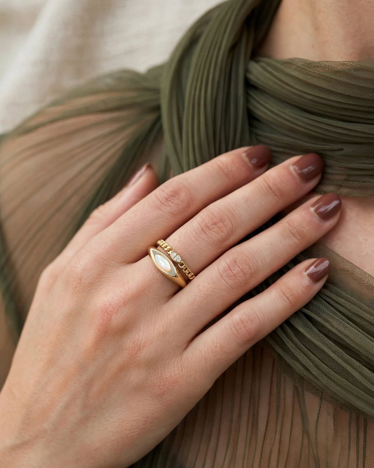 A close-up of a person's hand wearing a delicate gold ring with a white marquise-shaped stone and small clear stones, styled with an olive green pleated top.