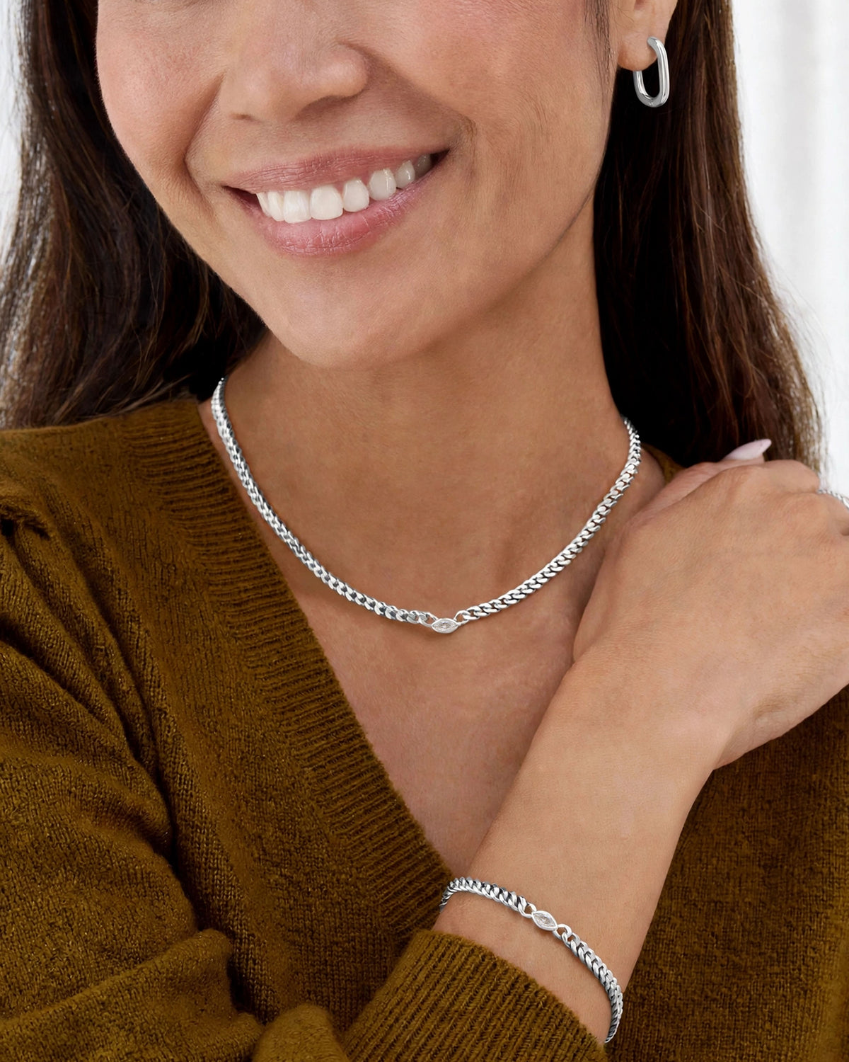 A close-up lifestyle shot of a smiling woman wearing a silver curb chain necklace and a matching silver cable bracelet, paired with a brown knit sweater.
