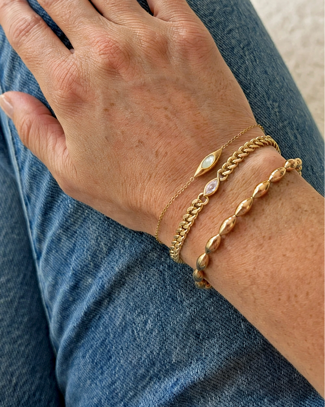 A close-up of a person's hand resting on their lap, wearing three delicate gold bracelets, including a chain bracelet with a small oval charm and a thin chain bracelet with a clear stone.